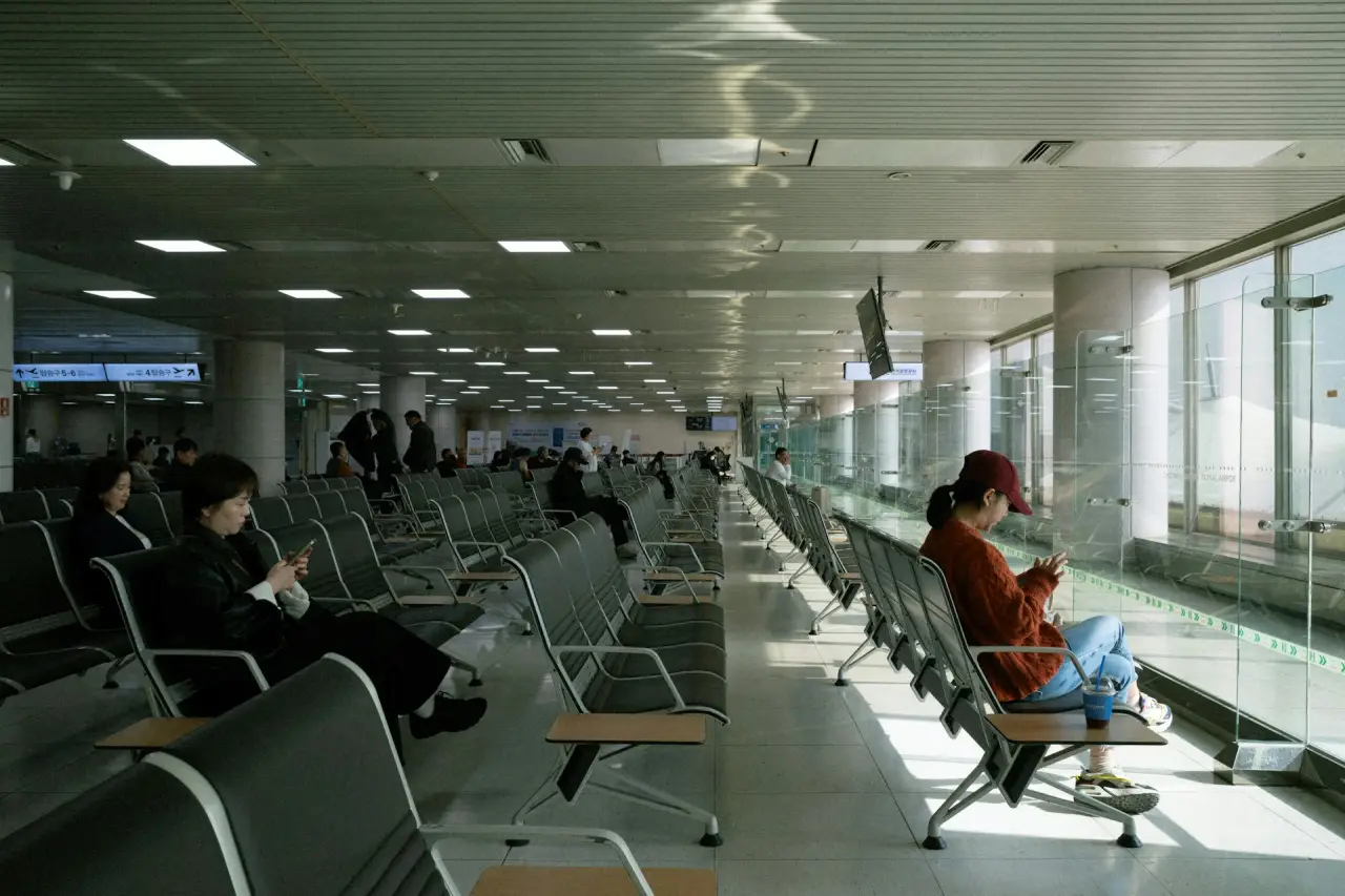 Travelers seated in an airport waiting area, checking their phones near large windows