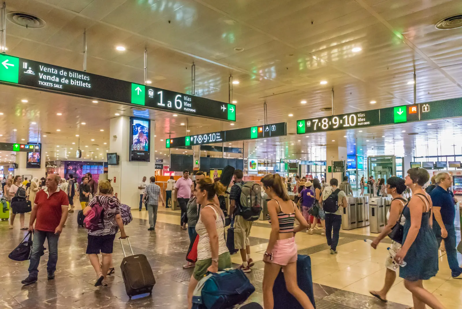 Travelers walking through Barcelona Sants railway station concourse with luggage.