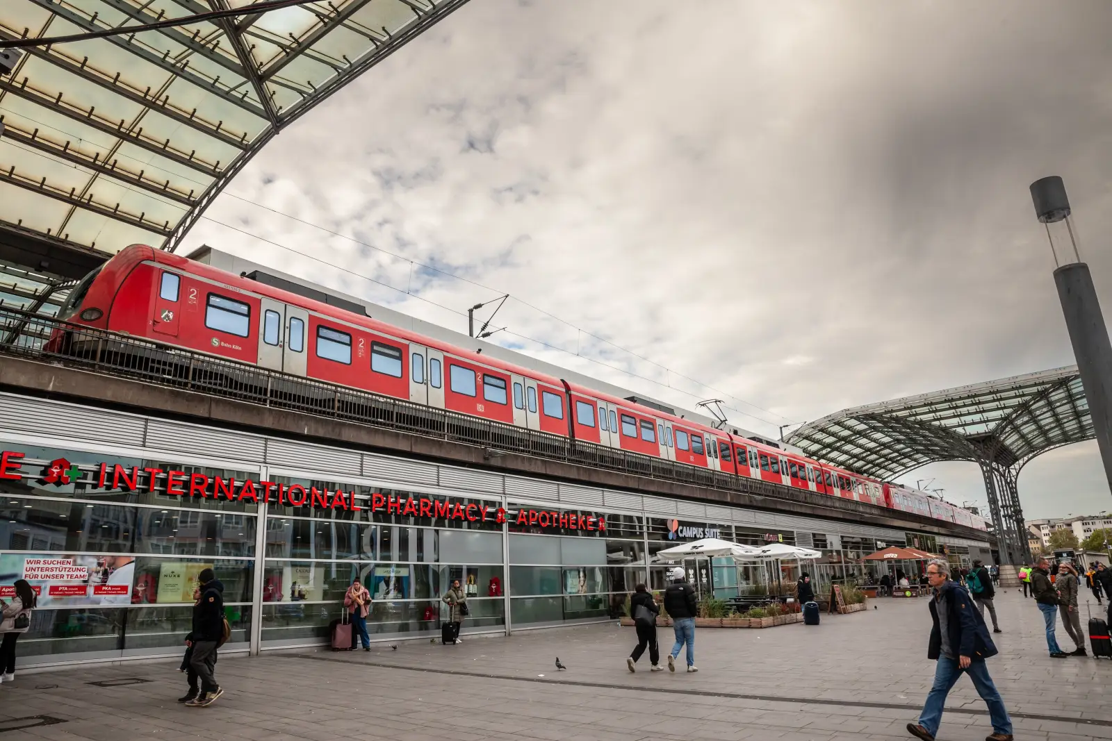 Red S-Bahn train arriving at Cologne Central Station (Köln Hauptbahnhof) in Germany, with people walking on the station plaza below.