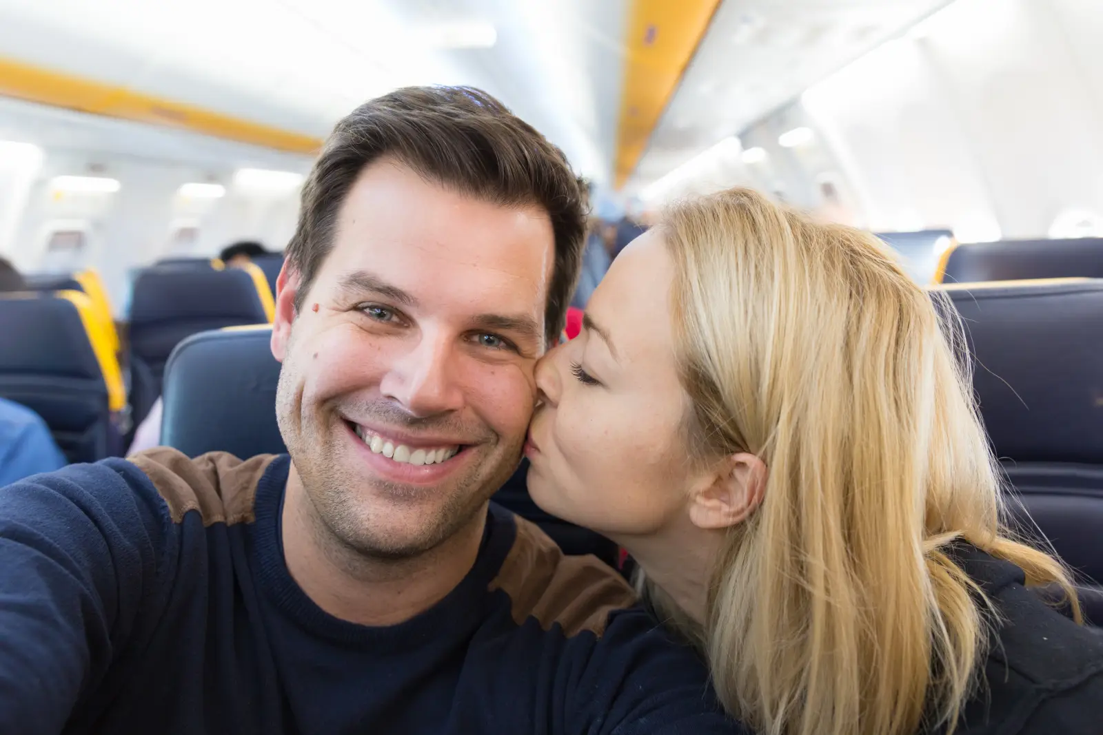 Couple sitting on an airplane seat, smiling as the woman kisses the man on the cheek