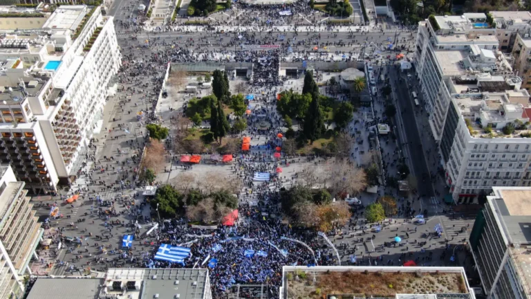 Large protest in central Athens with crowds carrying Greek flags and banners gathered in a city square.