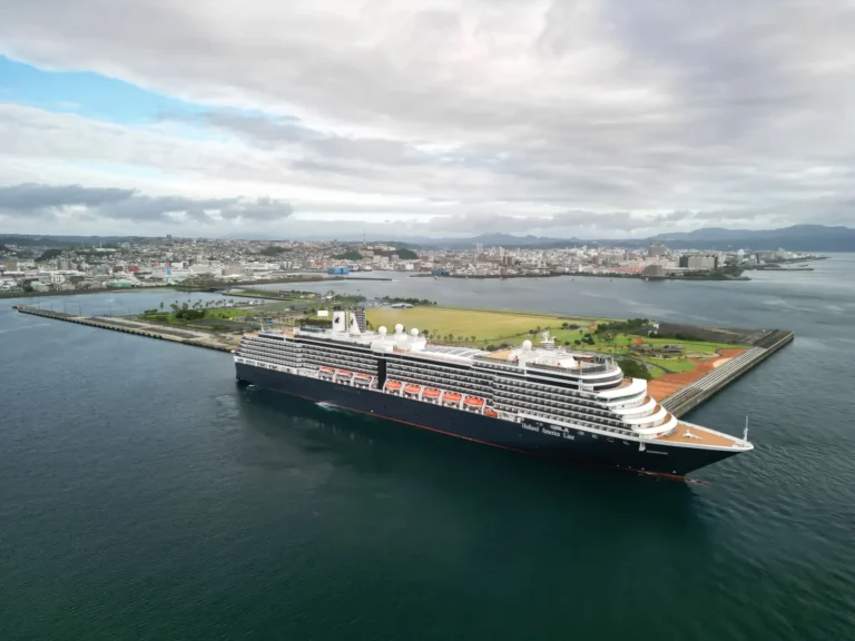 Holland America Line’s cruise ship Noordam docked at a Japanese port with a coastal city and mountains in the background.