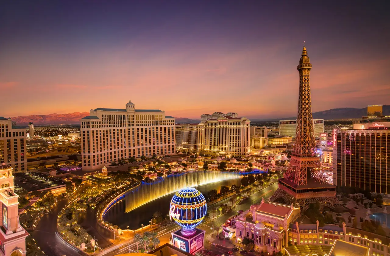View of the Las Vegas Strip at sunset with the Bellagio fountains, Paris Las Vegas Eiffel Tower replica, and Caesars Palace illuminated.