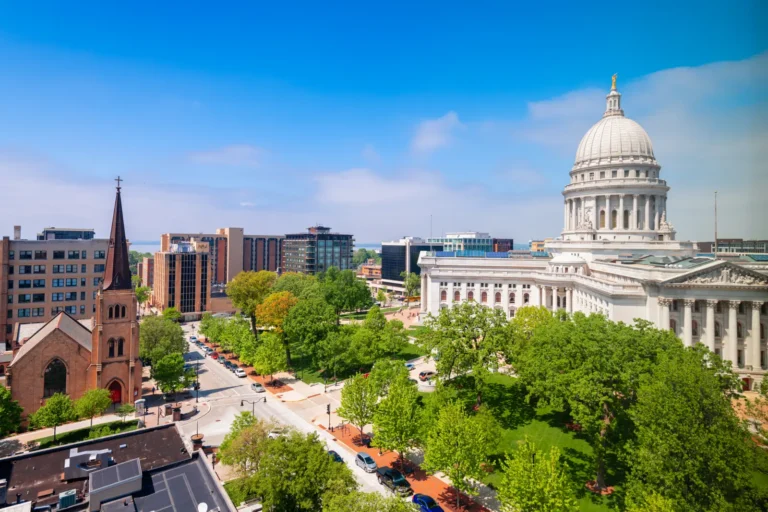 An aerial view of the Wisconsin State Capitol building in Madison with its white dome and columned facade, surrounded by lush green trees and park grounds, next to a tall, historic brick church spire. The background features city buildings and a bright blue sky.