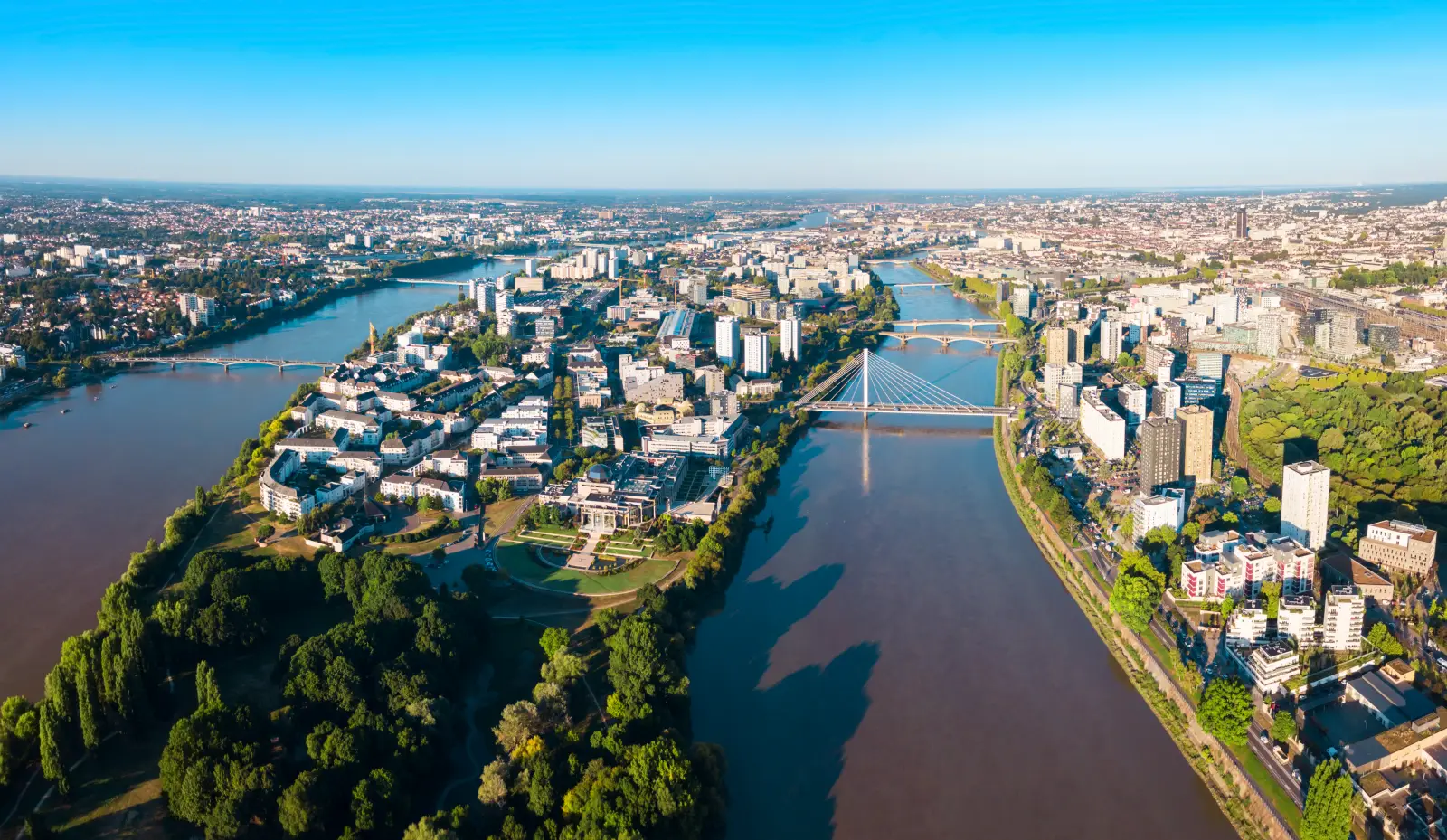 Aerial view of Nantes, France, showing the Loire River winding through the city with bridges, parks, and urban districts under clear skies.