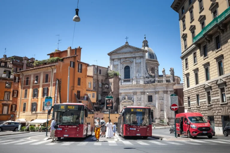 Two red ATAC city buses stopped in central Rome near a historic church.