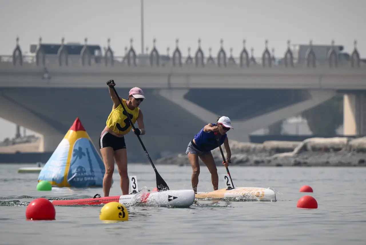 Abu Dhabi Turns Up the Heat for 2025 Stand Up Paddling World Championships 39 Two women compete in a stand up paddling race on calm waters in Abu Dhabi, with a bridge in the background.