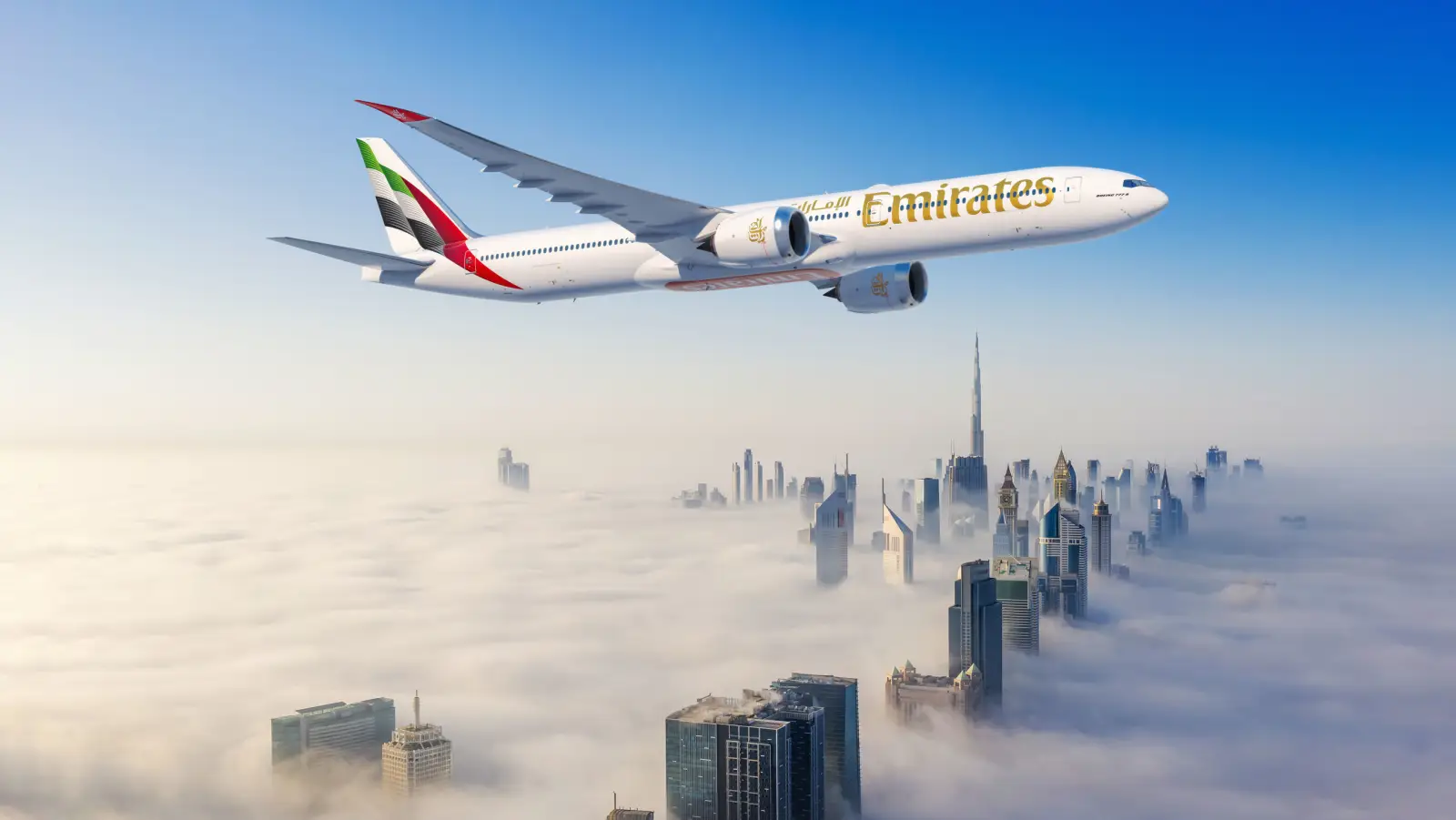 Emirates aircraft flying above fog-covered Dubai skyline with Burj Khalifa in view.