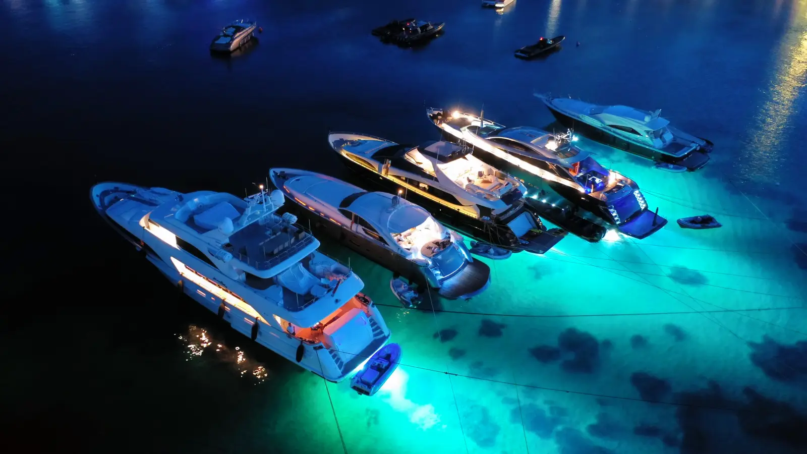 Luxury yachts anchored at night with glowing underwater lights illuminating turquoise water.