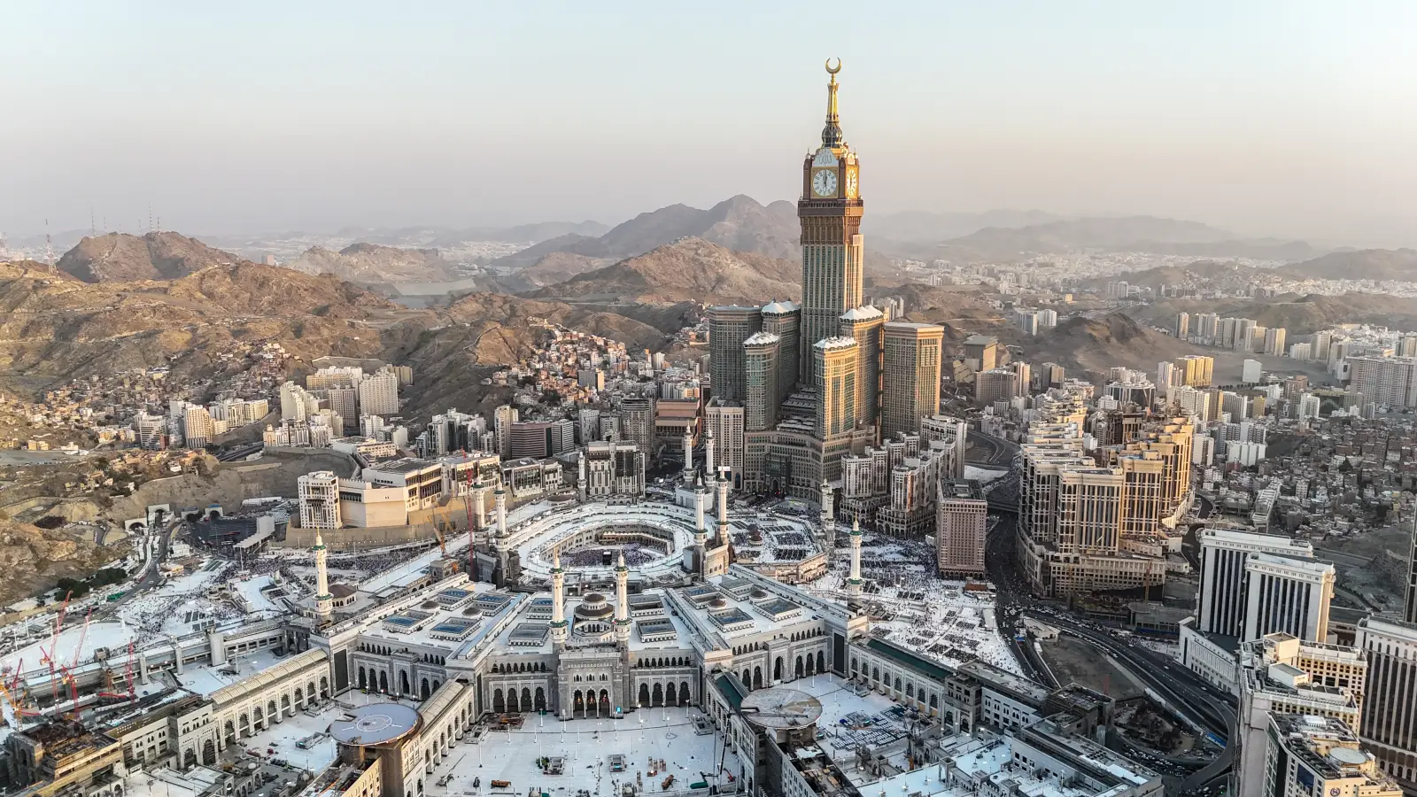 Aerial view of the Great Mosque and Abraj Al Bait Towers in Makkah surrounded by mountains at sunrise.