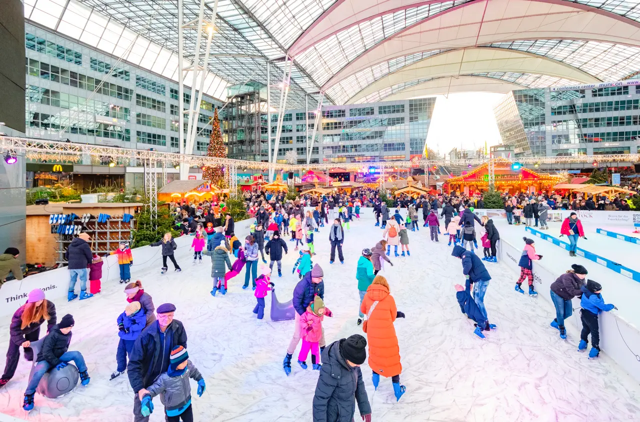Crowds ice skating under the glass roof at Munich Airport’s Christmas and winter market surrounded by festive stalls and lights.