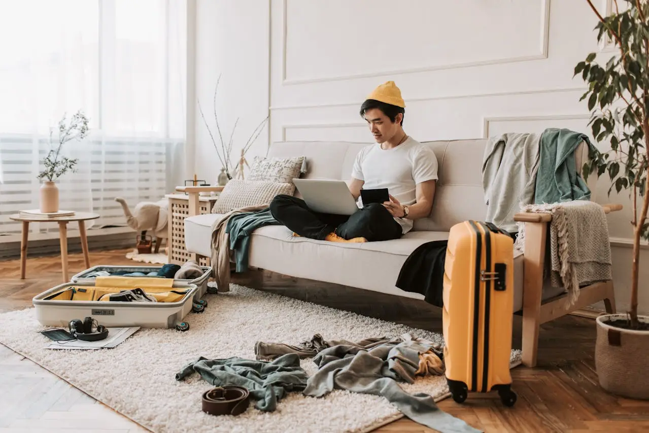 Person sitting on a sofa using a laptop while packing clothes and a yellow suitcase in a cozy room.
