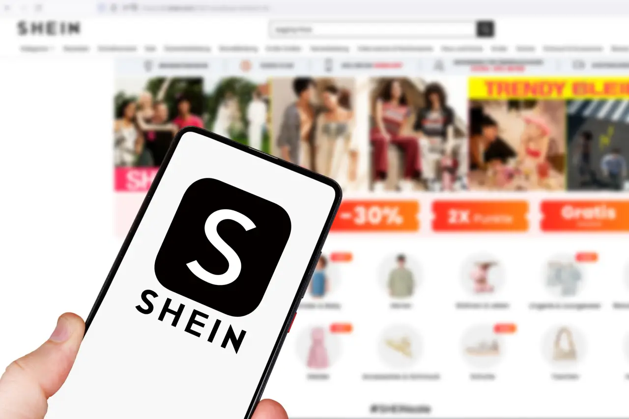 Person holding a smartphone displaying the Shein logo, with the Shein online store open in the background.