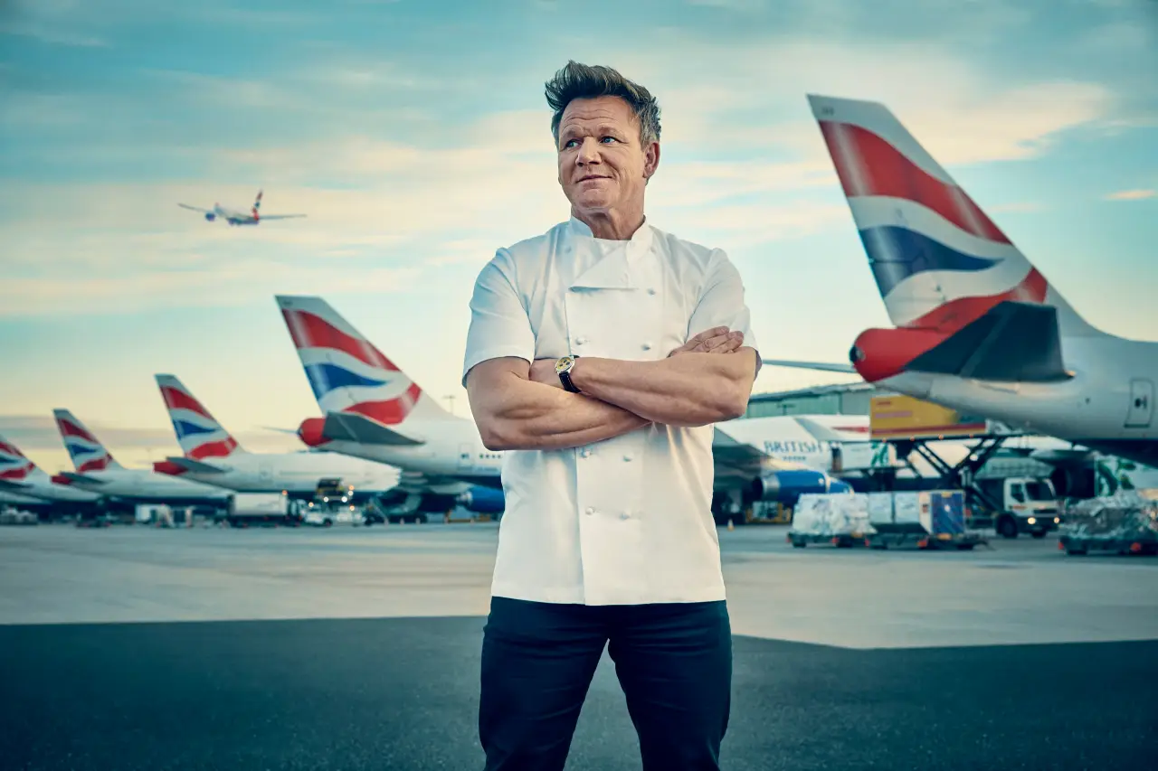 Gordon Ramsay Chef in white jacket stands on airport tarmac with aircraft in the background