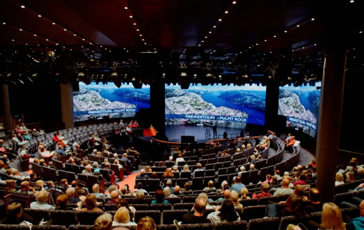 Audience attending a travel presentation in a cruise ship theater with large scenic screens