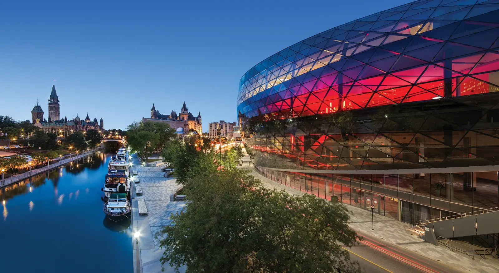 Rideau Canal at dusk with boats moored along the waterfront and the illuminated glass façade of the Shaw Centre in Ottawa.