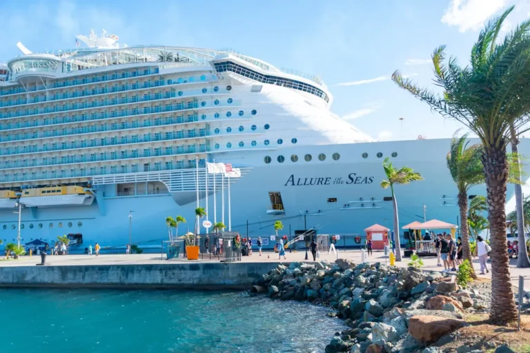 Royal Caribbean’s Allure of the Seas docked at a sunny waterfront port as passengers explore the pier lined with palm trees.