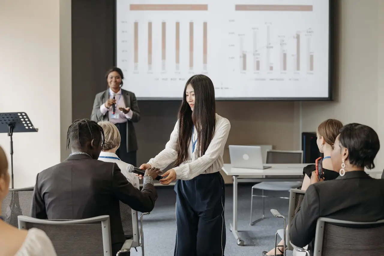 Conference staff member hands a microphone to an attendee during a business presentation with charts displayed on a screen.