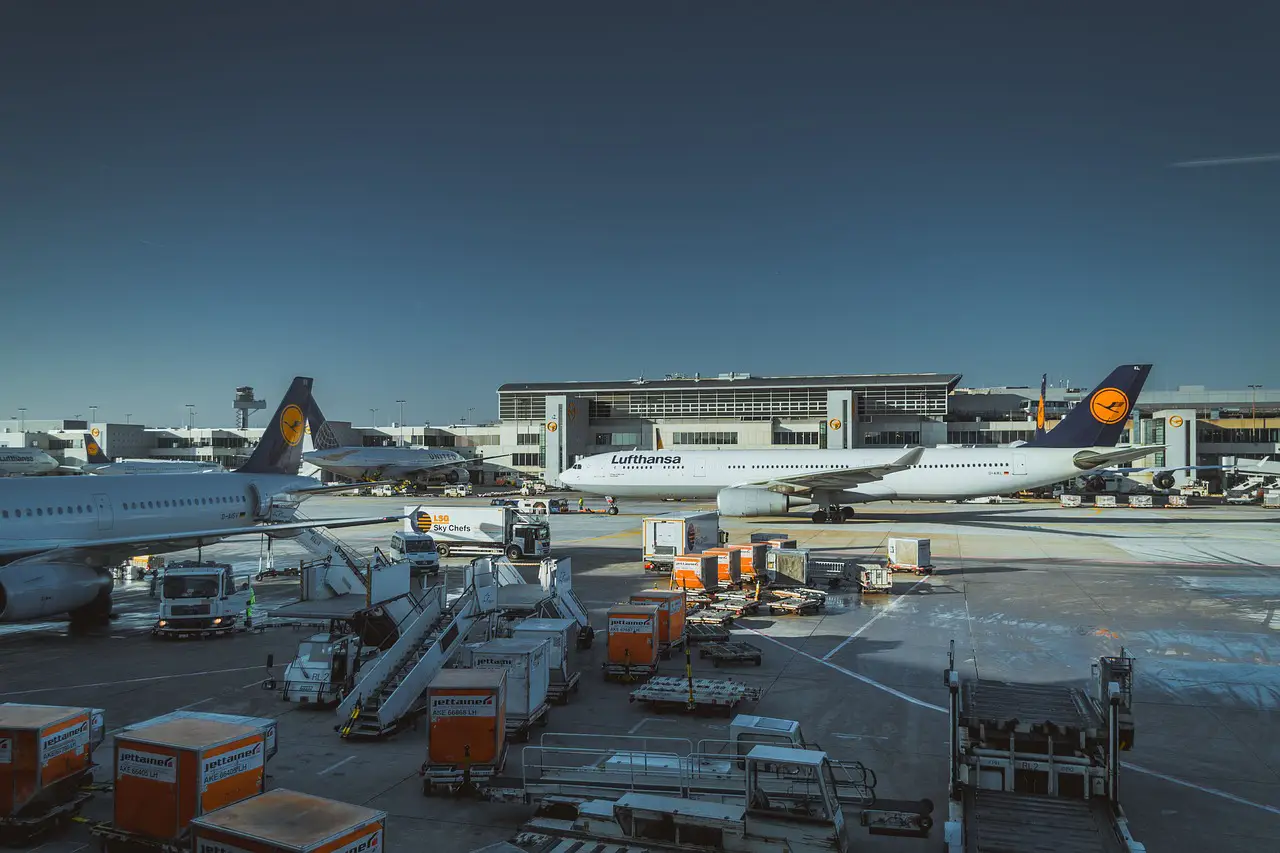Airport apron scene with multiple Lufthansa aircraft parked at gates, surrounded by ground service vehicles, catering trucks, loading equipment, and cargo containers.