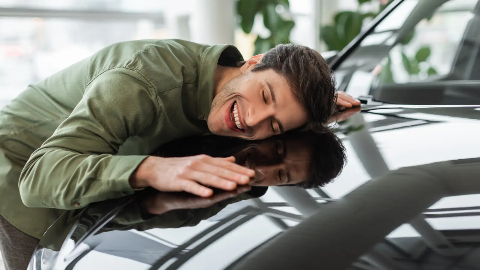 Smiling man hugging the hood of a shiny black car inside a showroom.