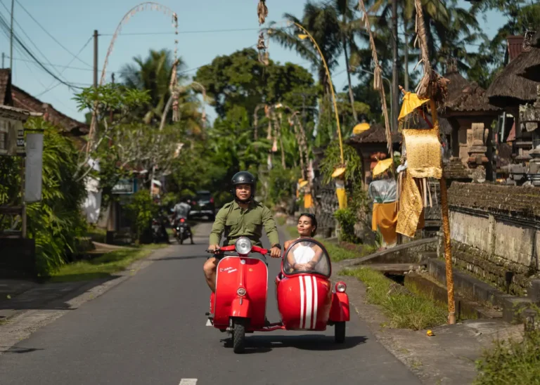 Other News 13 A person rides a red scooter with a sidecar through a lush village road in Bali, while the passenger relaxes and enjoys the scenery.