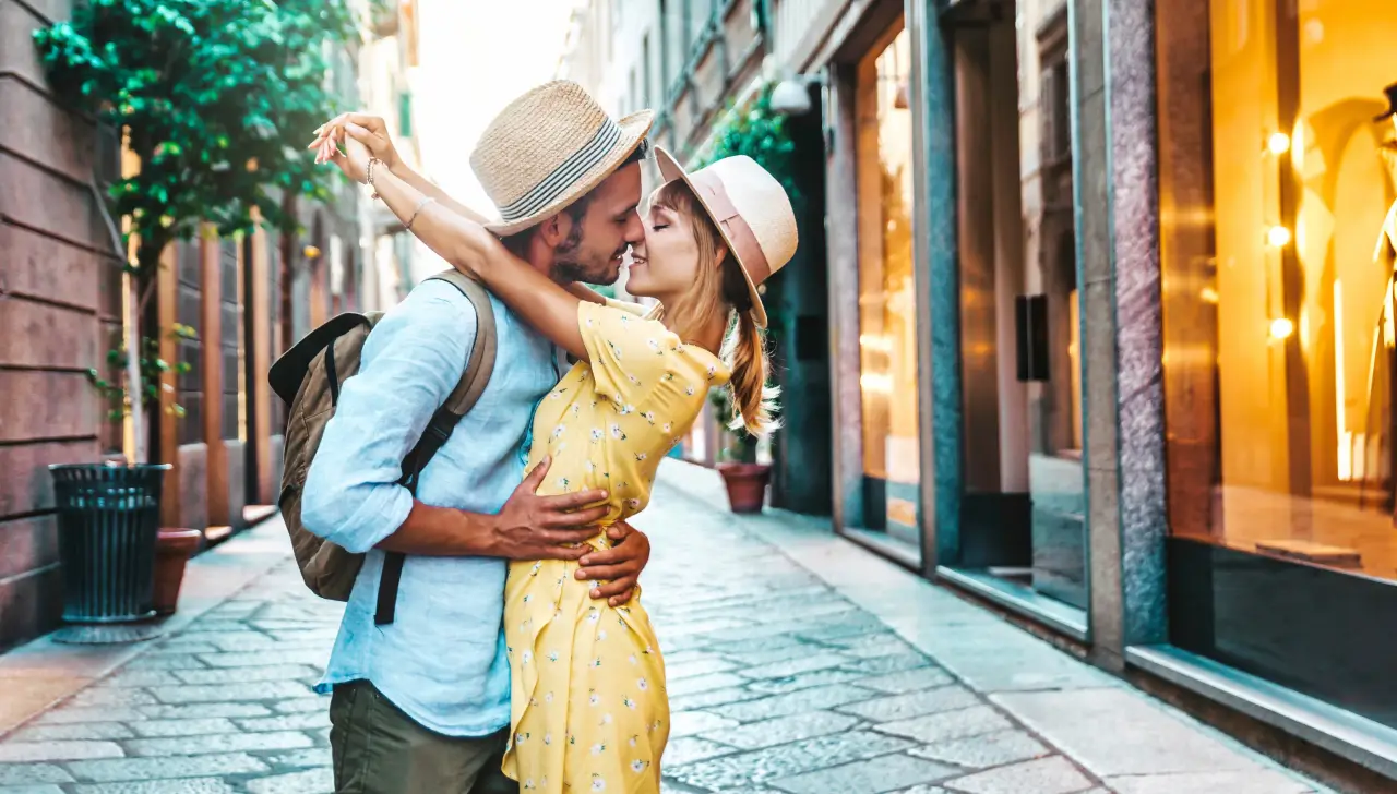 Couple kissing in a narrow European street, wearing summer clothes and straw hats