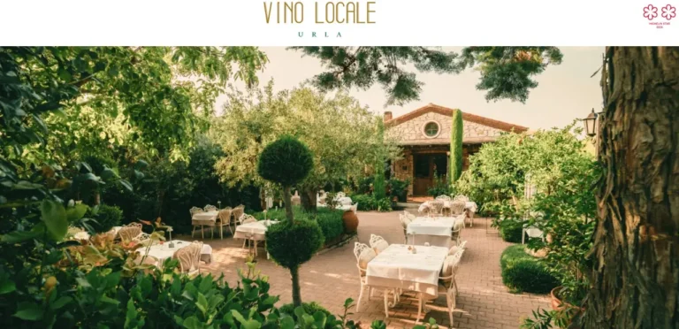 Outdoor dining area at Vino Locale in Urla, Turkey, featuring white table settings surrounded by lush greenery and a stone villa-style building in the background.