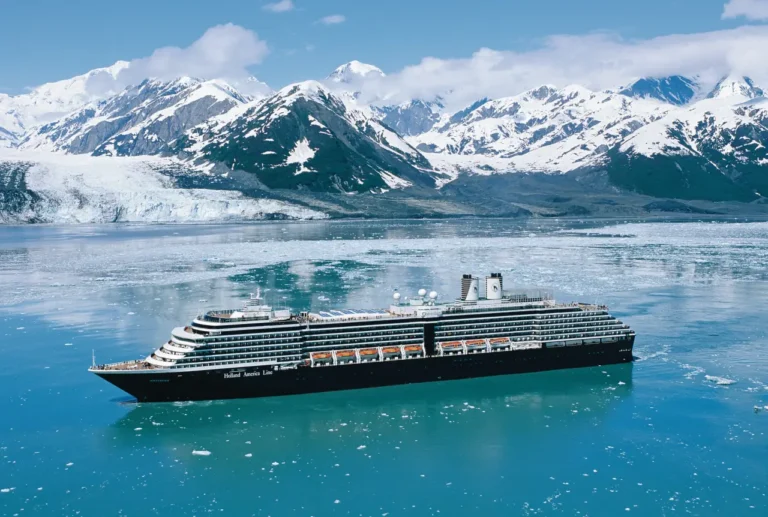 Holland America Line cruise ship sailing past a glacier in Alaska, surrounded by icy blue water and snow-capped mountains.