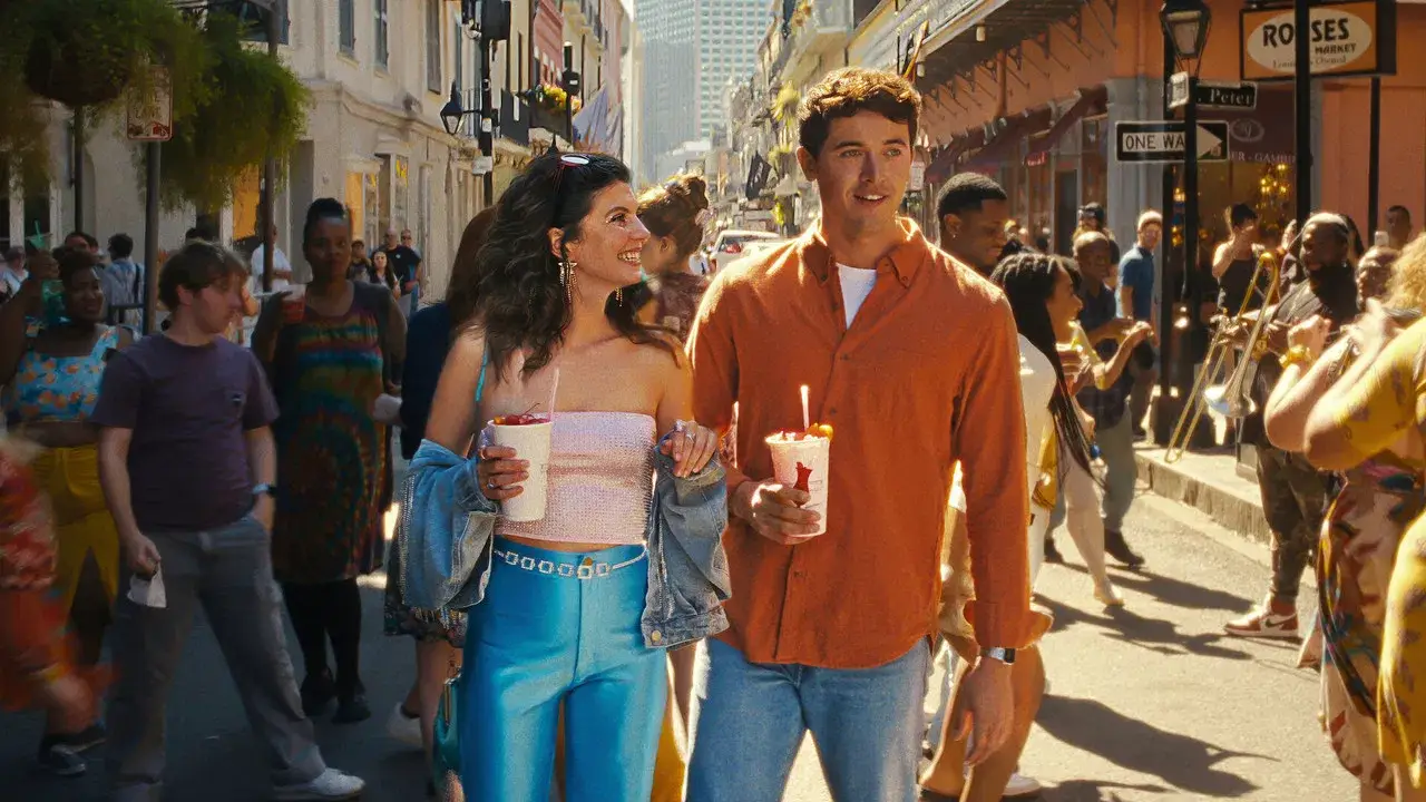 A smiling couple walks through a lively street festival, holding drinks as musicians and crowds fill the sunlit background.