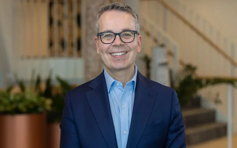 Smiling business professional wearing glasses and a navy suit, standing indoors with a softly blurred staircase and plants in the background.