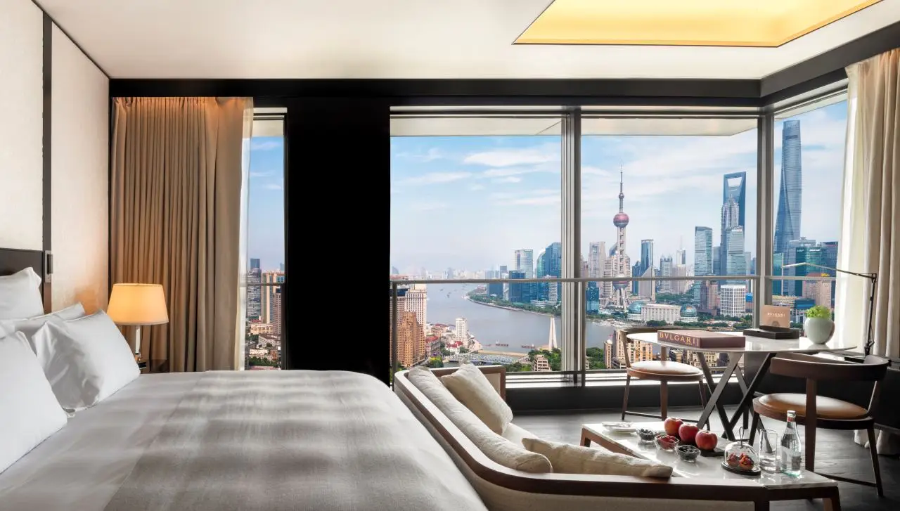 Shanghai, London And Dubai Lead In New Hotel Room Openings For 2026 3 Luxury hotel room interior with floor-to-ceiling windows overlooking the Shanghai skyline and Huangpu River, featuring a modern bed, seating area, and city view.