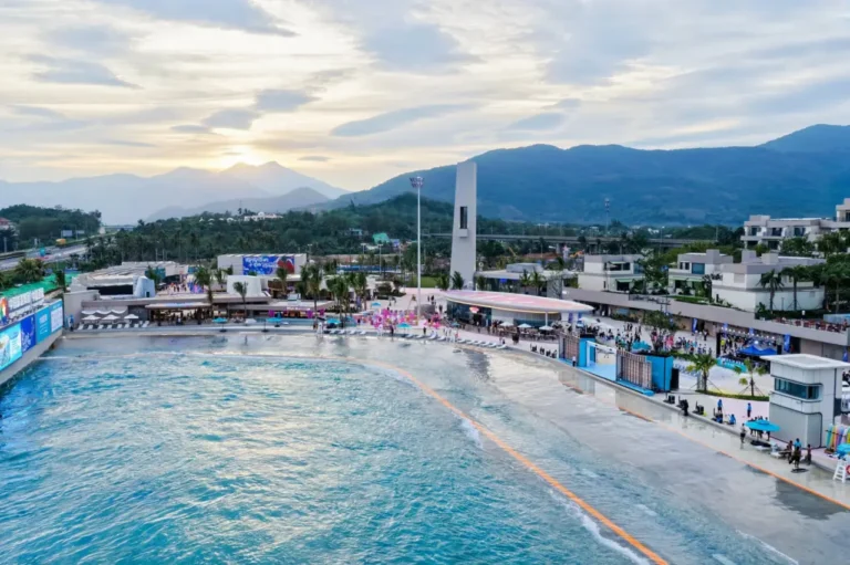 Aerial view of a large artificial surf lagoon at a coastal resort, with mountains in the background, sunset sky, and visitors along the shoreline.