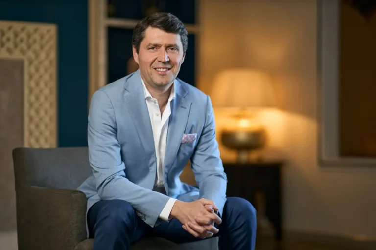 Portrait of Thomas Krooswijk seated indoors, wearing a light blue blazer and smiling at the camera.