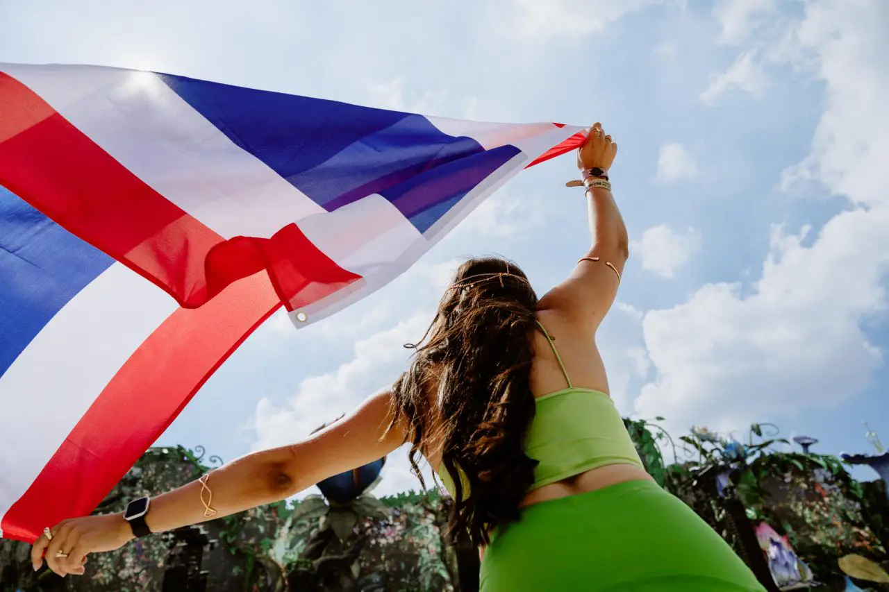 Pattaya In Thailand Chosen To Host Electronic Dance Festival Tomorrowland, Its First Outdoor Edition In Asia 3 Festivalgoer waving a Thai flag above a crowd at an outdoor music festival under a bright blue sky.