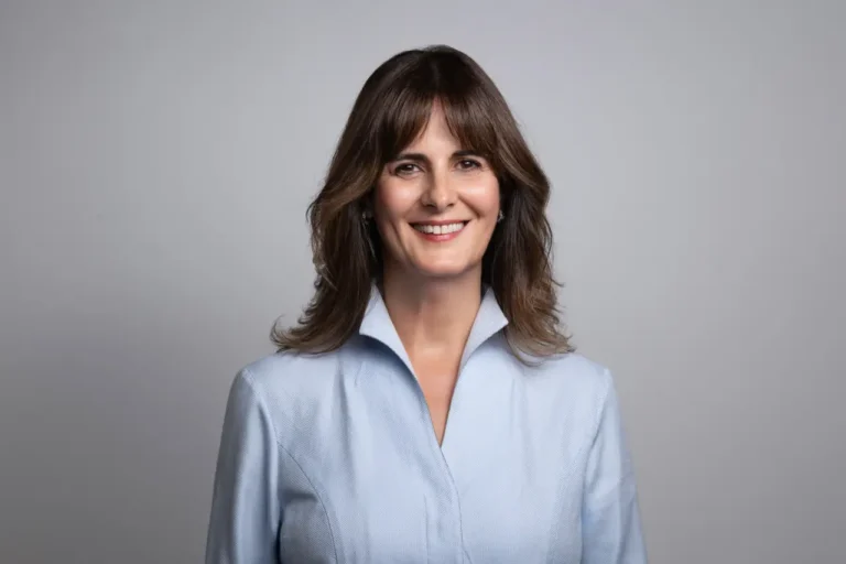 Smiling woman; Yeşim Gürer Oymak in a light blue jacket posing for a professional studio portrait against a neutral background