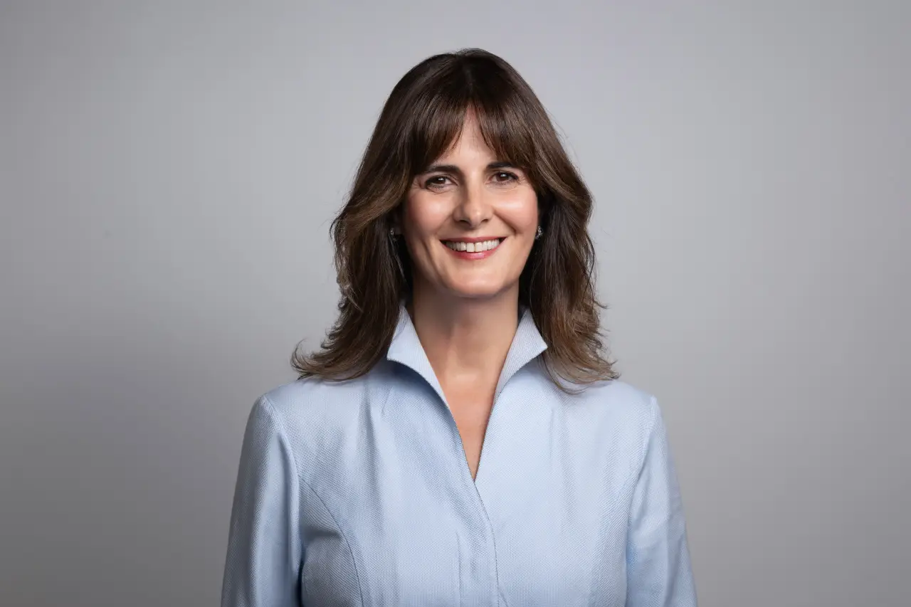 Smiling woman; Yeşim Gürer Oymak in a light blue jacket posing for a professional studio portrait against a neutral background