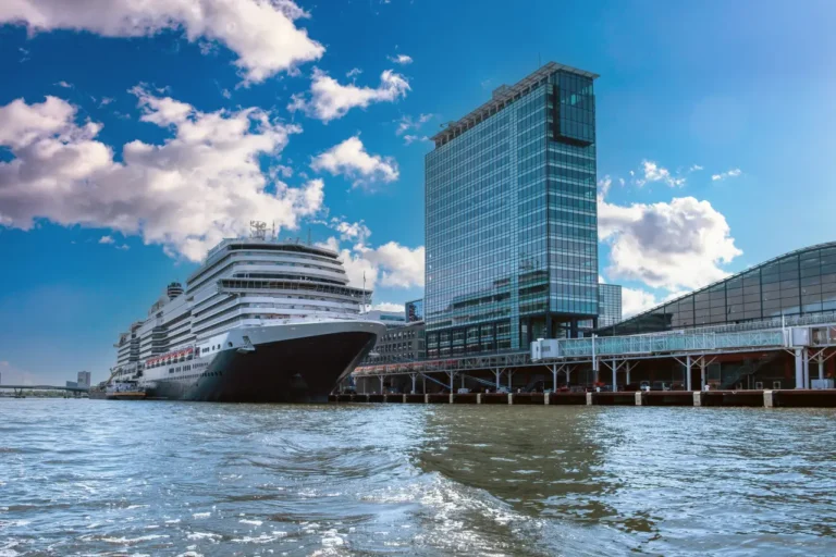 Luxury cruise ship docked at Amsterdam harbor terminal beside modern glass office buildings under a blue, partly cloudy sky.