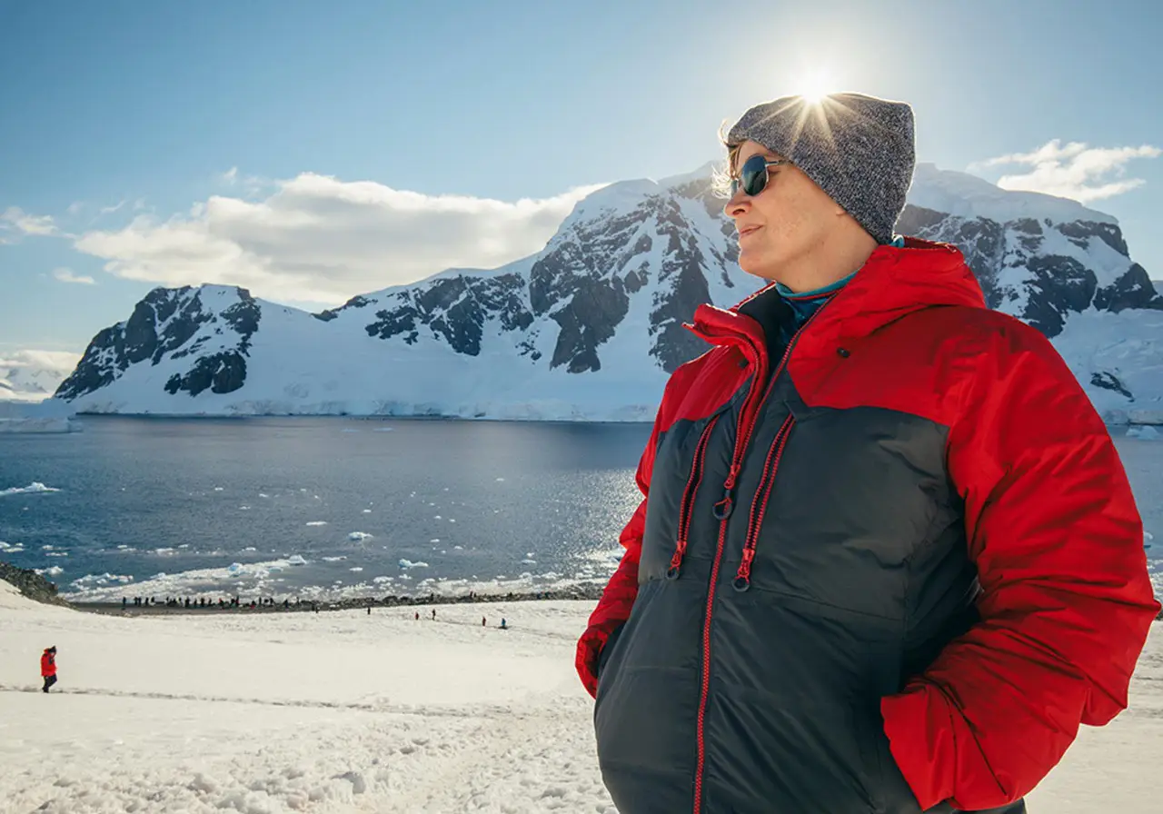 Tourist in a red winter jacket standing on a snowy Antarctic coastline with icebergs and mountains in the background