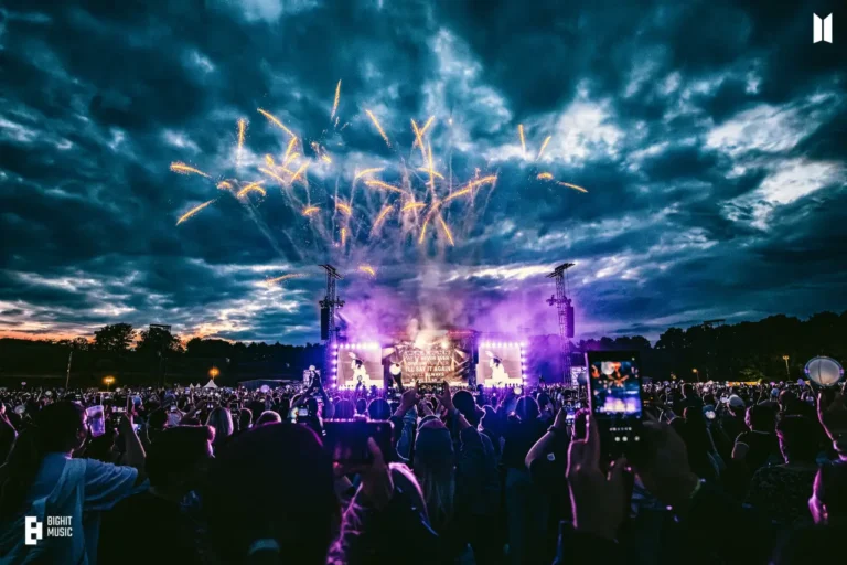 Fireworks explode above a packed outdoor BTS concert stage at night, with fans holding glowing light sticks and smartphones.