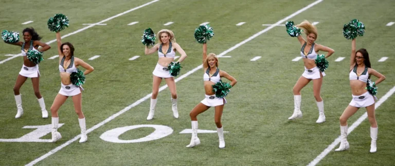 Cheerleaders perform with green pom-poms on a football field during a game.