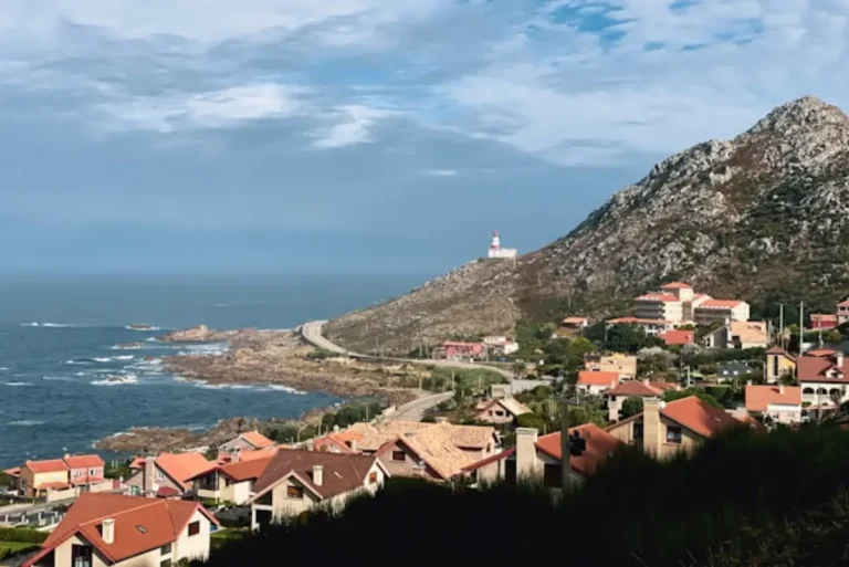 Spain Travel And Tourism News 23 Coastal village with red-roof houses, rocky shoreline, winding road, and a lighthouse on a hillside overlooking the ocean.