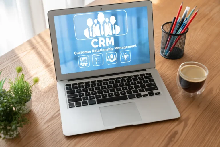 Laptop on a wooden desk displaying a CRM dashboard with customer relationship management icons, beside a coffee cup, plant and stationery.