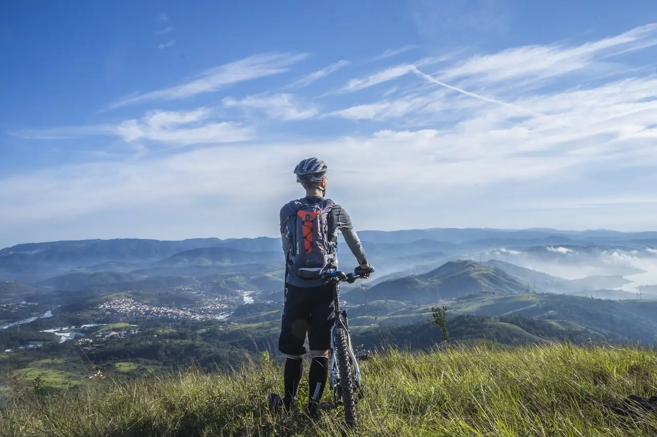 Cyclist stands on a grassy mountain viewpoint overlooking rolling hills, valleys and distant villages under a clear blue sky.