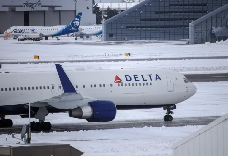 A Delta Air Lines aircraft taxis on a snow-covered runway at an airport, with other planes and hangars in the background.