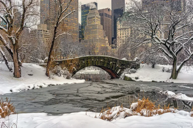 Snow-covered Gapstow Bridge arches over a partially frozen pond in Central Park, with Manhattan skyscrapers in the background at early morning.