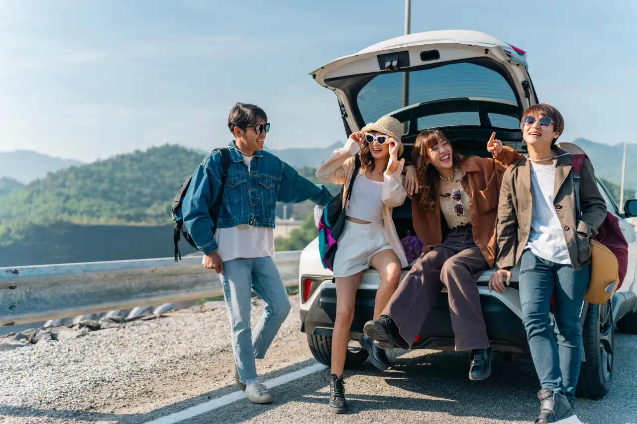 Gen Z Travel Is Changing Fast — And They Want Less Tech Telling Them What To Do 7 Group of young friends relaxing on the open trunk of an electric car during a summer road trip, enjoying mountain countryside views and an outdoor lifestyle.