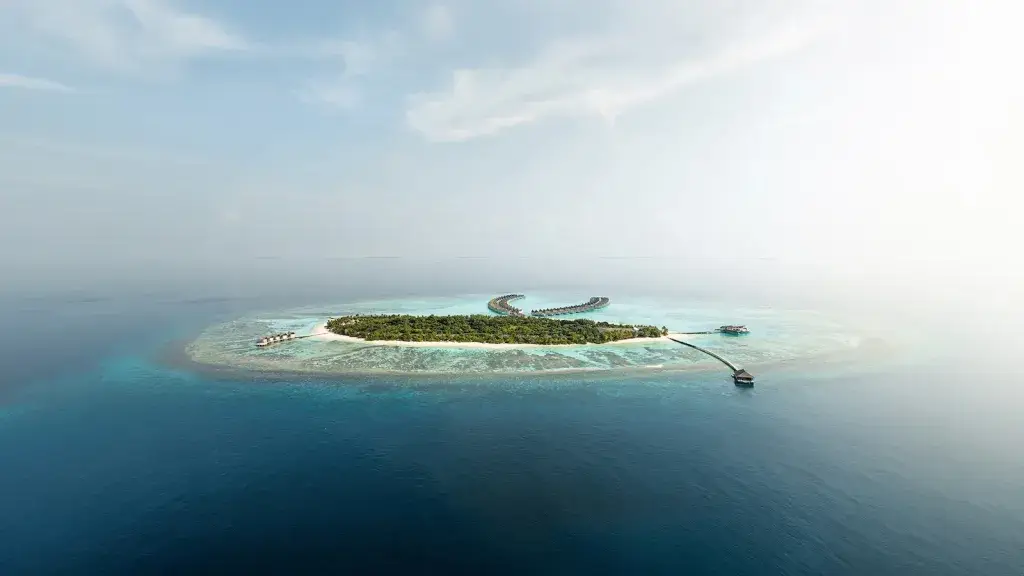 New Maldives Hotels &Amp; Resorts, 2025 &Amp; 2026 11 Aerial view of Mondrian Maldives island with curved overwater villas, surrounding reef, and deep blue ocean in Noonu Atoll.