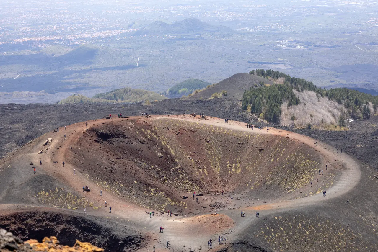 Visitors walk along the rim of a volcanic crater on Mount Etna, overlooking lava fields and distant towns in Sicily.