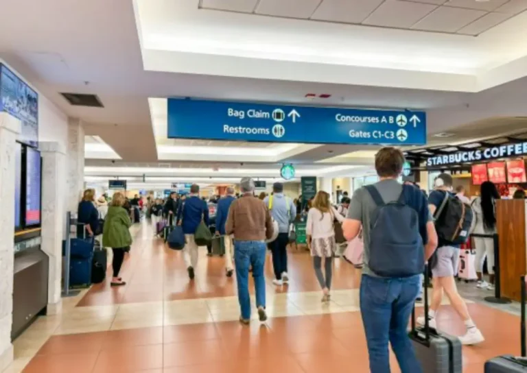 Passengers walk through a busy terminal at Palm Beach International Airport with overhead signs for bag claim, restrooms and concourses.
