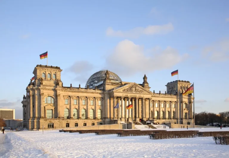 Snow-covered Reichstag building in Berlin with German flags flying under a clear winter sky.
